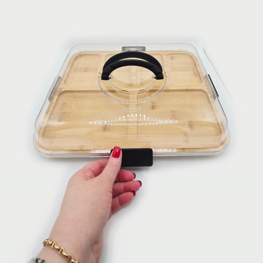 My hand on one of the latches of my bamboo charcuterie board with the clear lid closed and the handle up